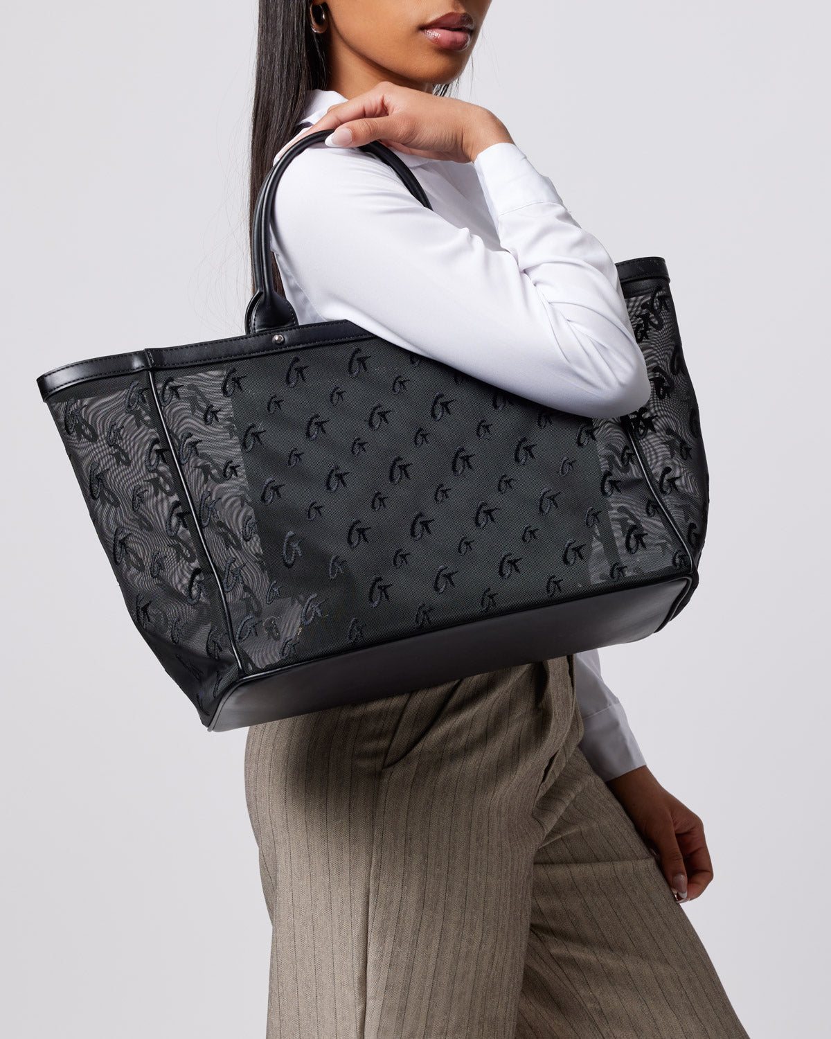 Black mesh monogram tote bag with logo pattern, held by a woman wearing a white shirt and beige pants.