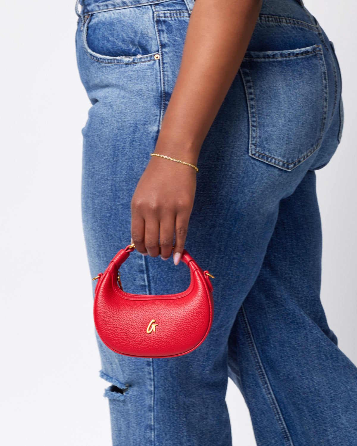 Red Nano Pebble Hobo Bag with gold logo, held by a person wearing blue jeans and a gold bracelet.