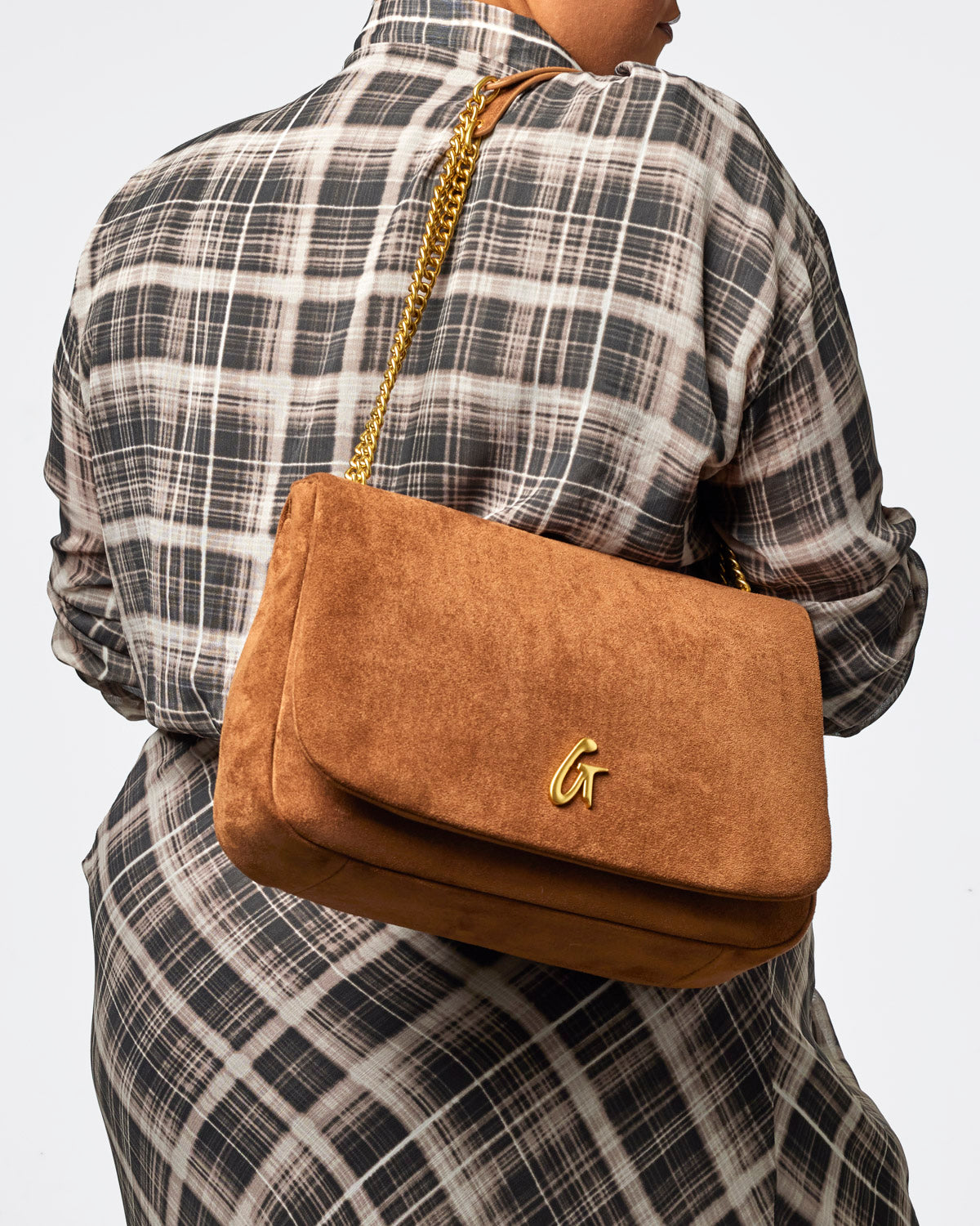 Medium brown suede flap bag with a gold chain strap, worn over the shoulder against a plaid shirt backdrop.