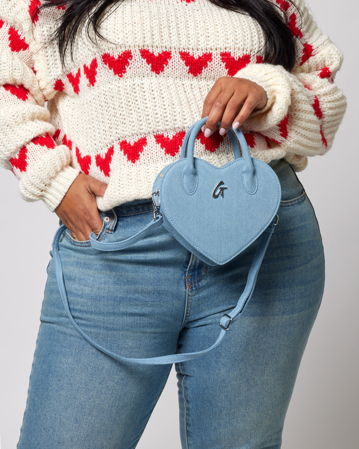 Light blue denim heart-shaped bag with a small "G" logo, carried by a woman in a cream and red heart-patterned sweater.