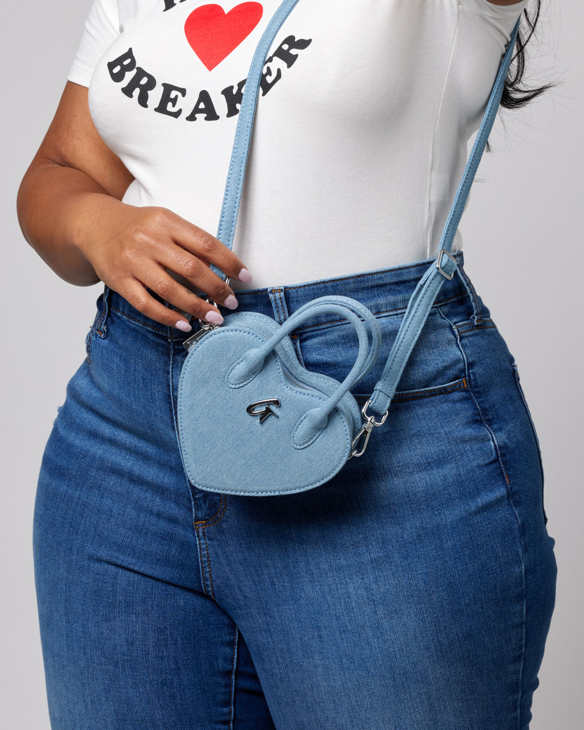 Small light blue denim heart-shaped bag with a strap, metal logo, and bow detail, worn crossbody style.