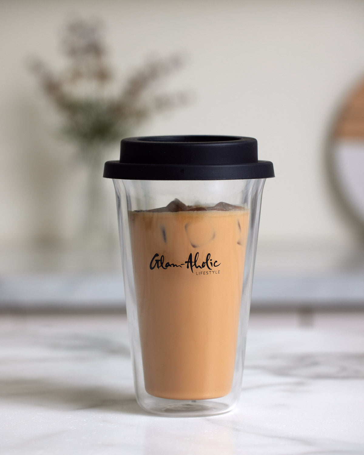 Clear double-walled tumbler with black lid, featuring "Glam-Aholic Lifestyle" logo, filled with iced coffee on a white surface.