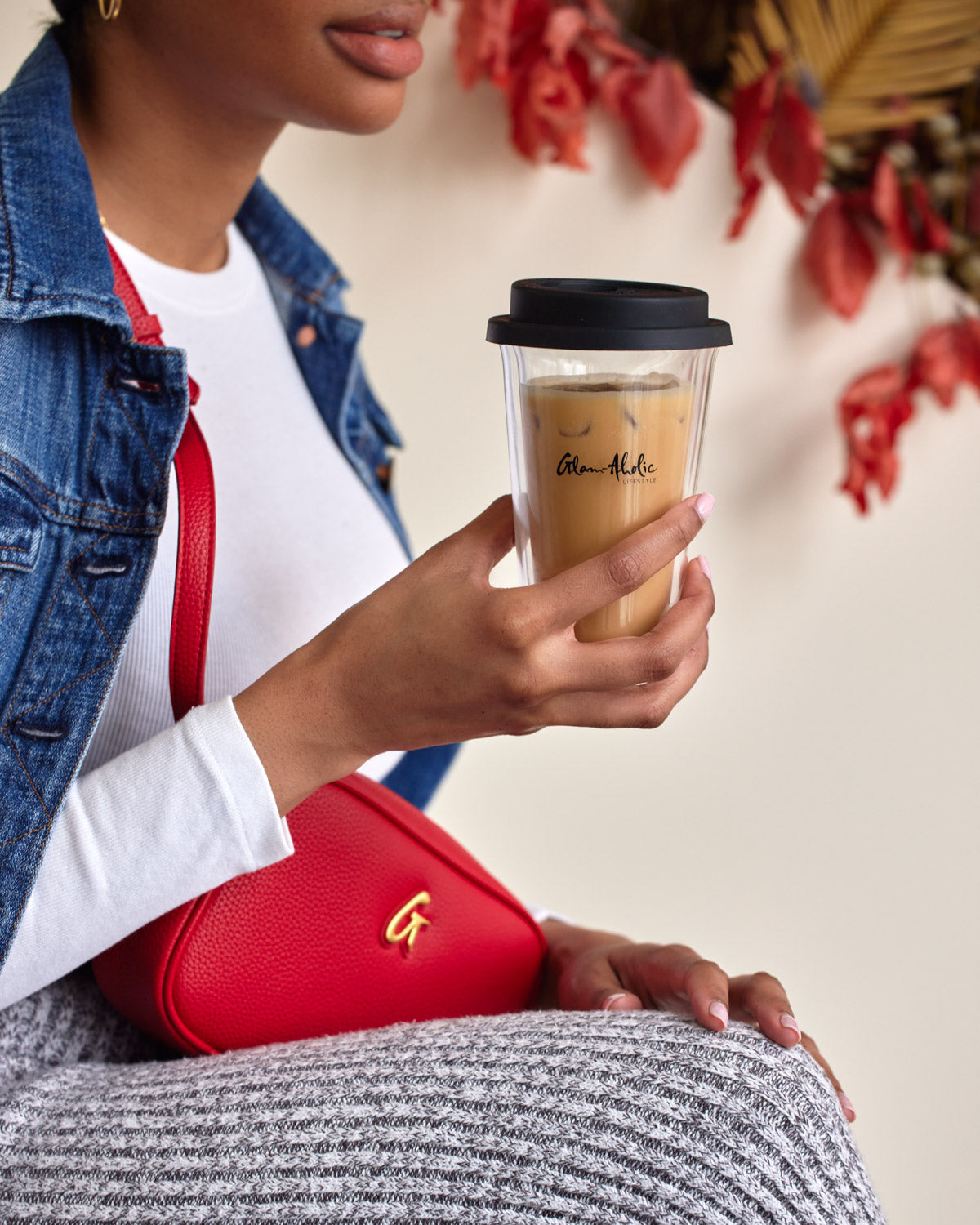 Close-up of a woman holding a signature logo tumbler with iced coffee, wearing a denim jacket and red purse.