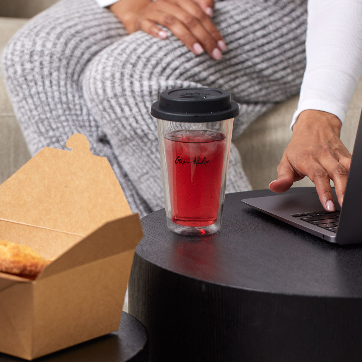 A clear tumbler with a black lid, featuring a signature logo, filled with red beverage and ice, placed on a black table.