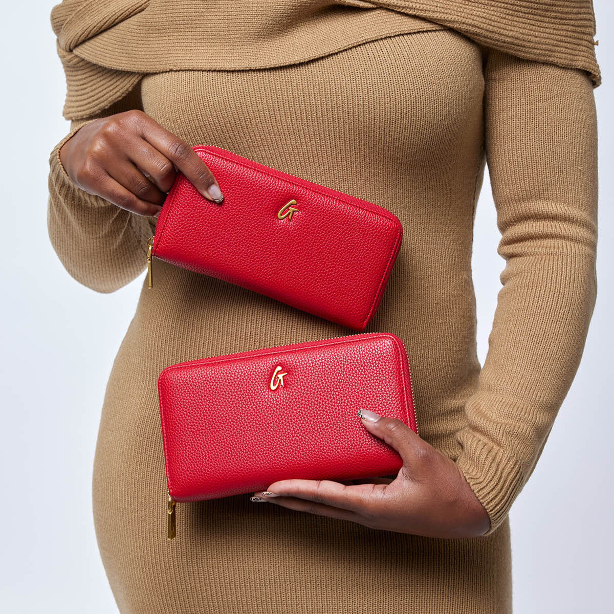 Red textured leather wallets with gold "G" logo, held by a woman in a tan sweater. Affordable luxury accessories for women.