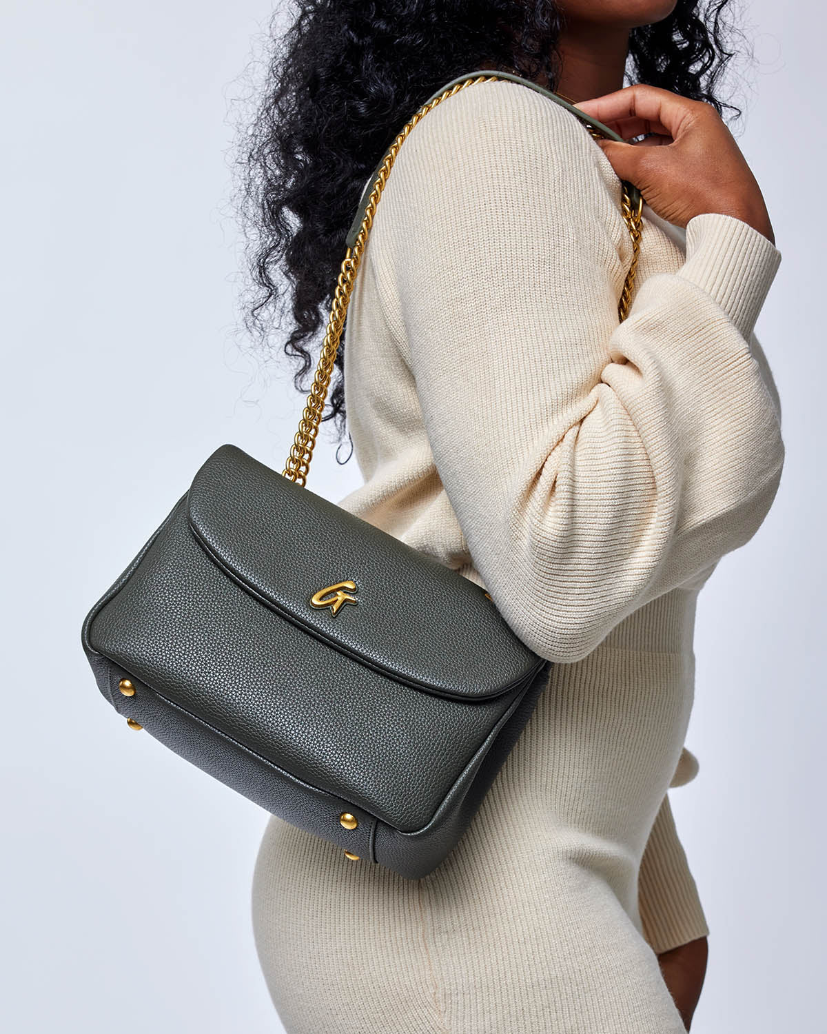 Medium olive green pebble leather flap bag with gold chain strap and logo, worn over a cream sweater by a woman.