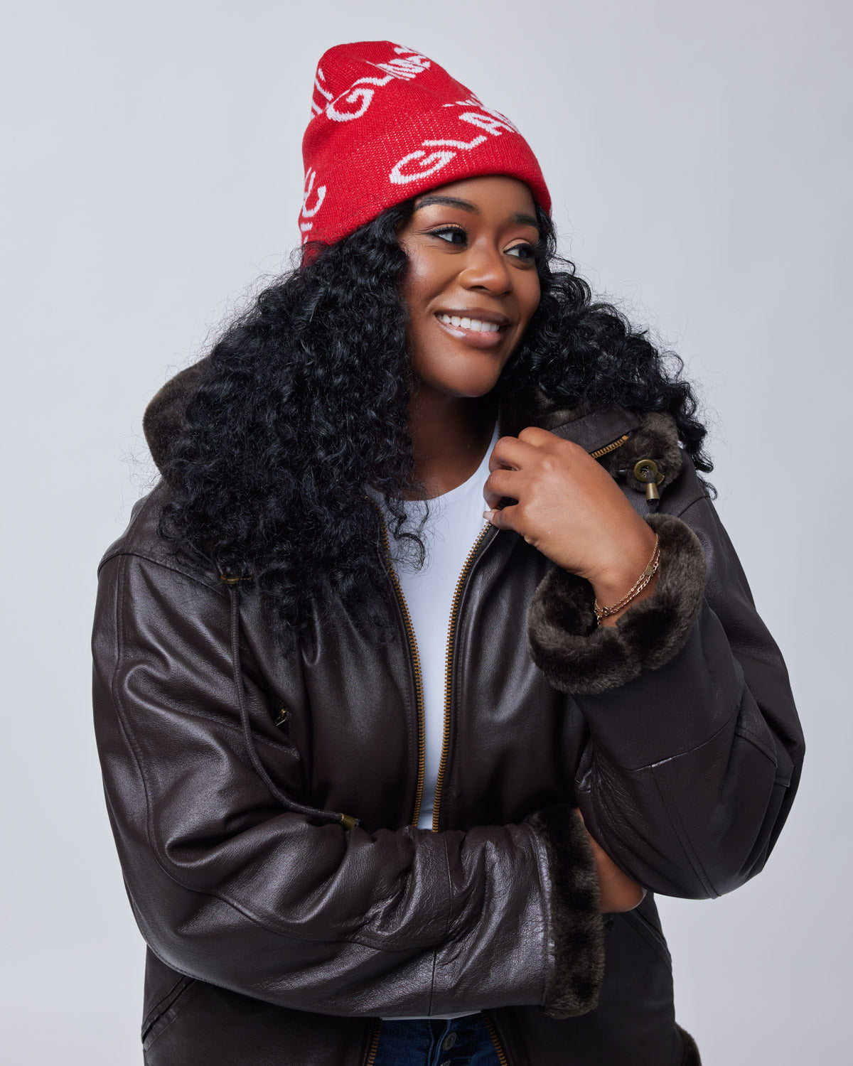 Woman wearing a red monogram beanie, dark brown leather jacket with fur cuffs, smiling against a light gray background.