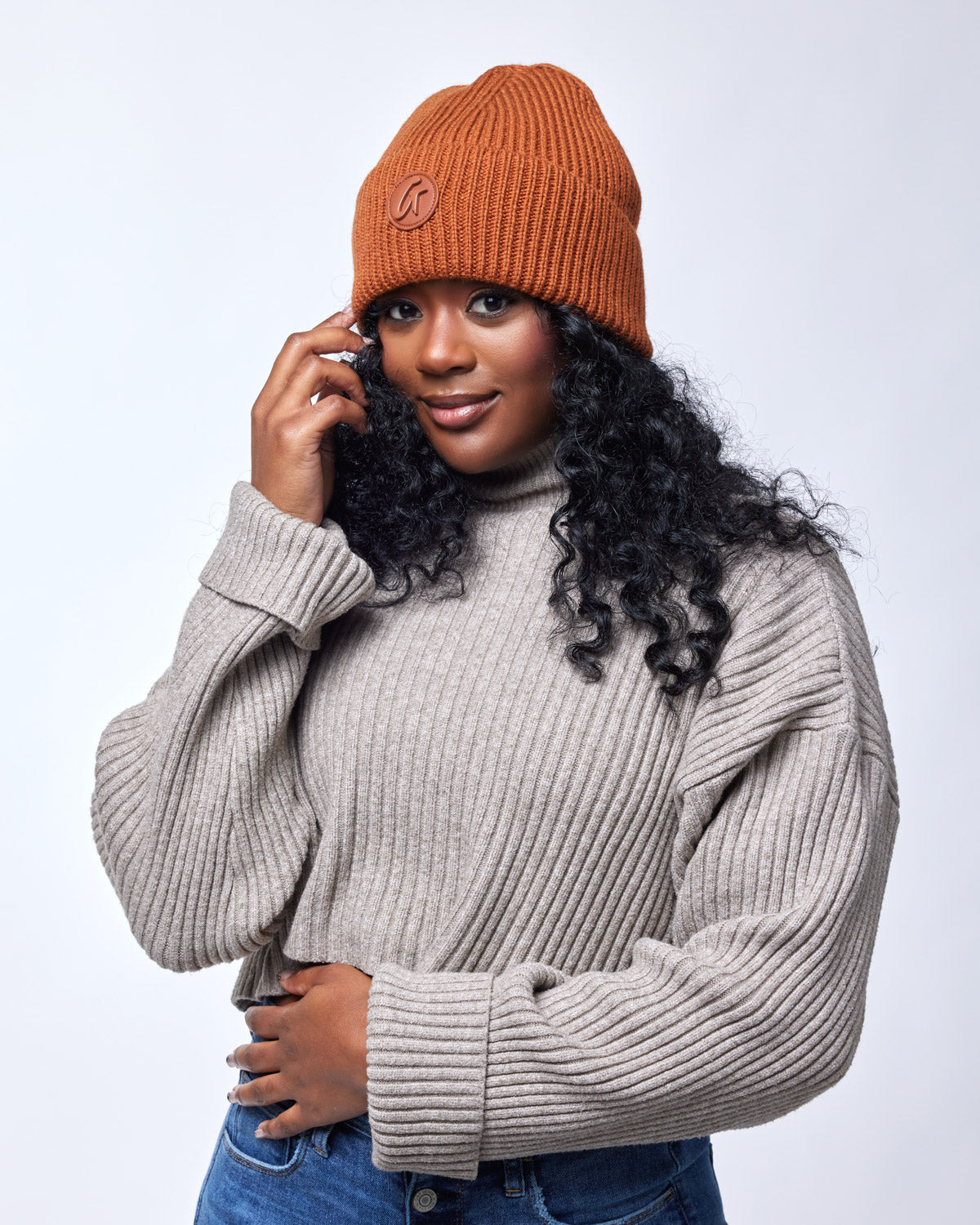 Woman wearing a brown knit beanie, beige ribbed sweater, and blue jeans, smiling softly against a plain background.
