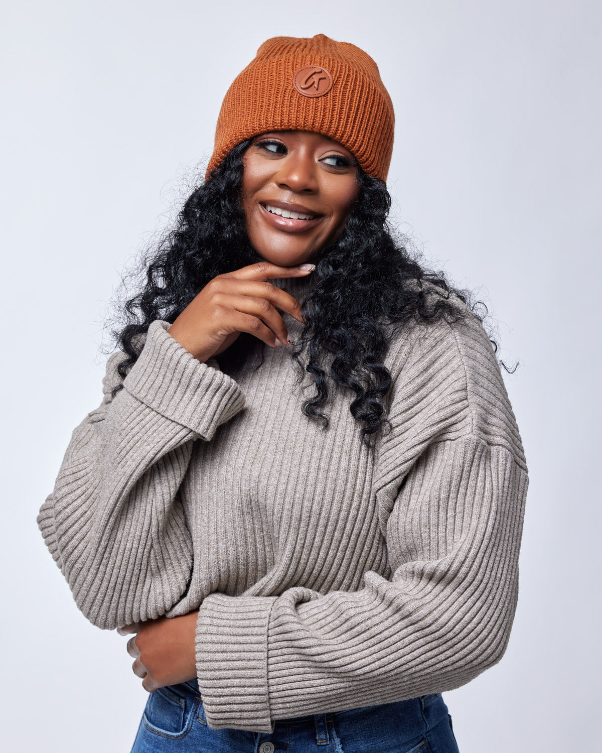 A woman wearing a brown knit beanie with a logo patch, smiling and posing with her hand near her chin, dressed in a beige ribbed sweater.
