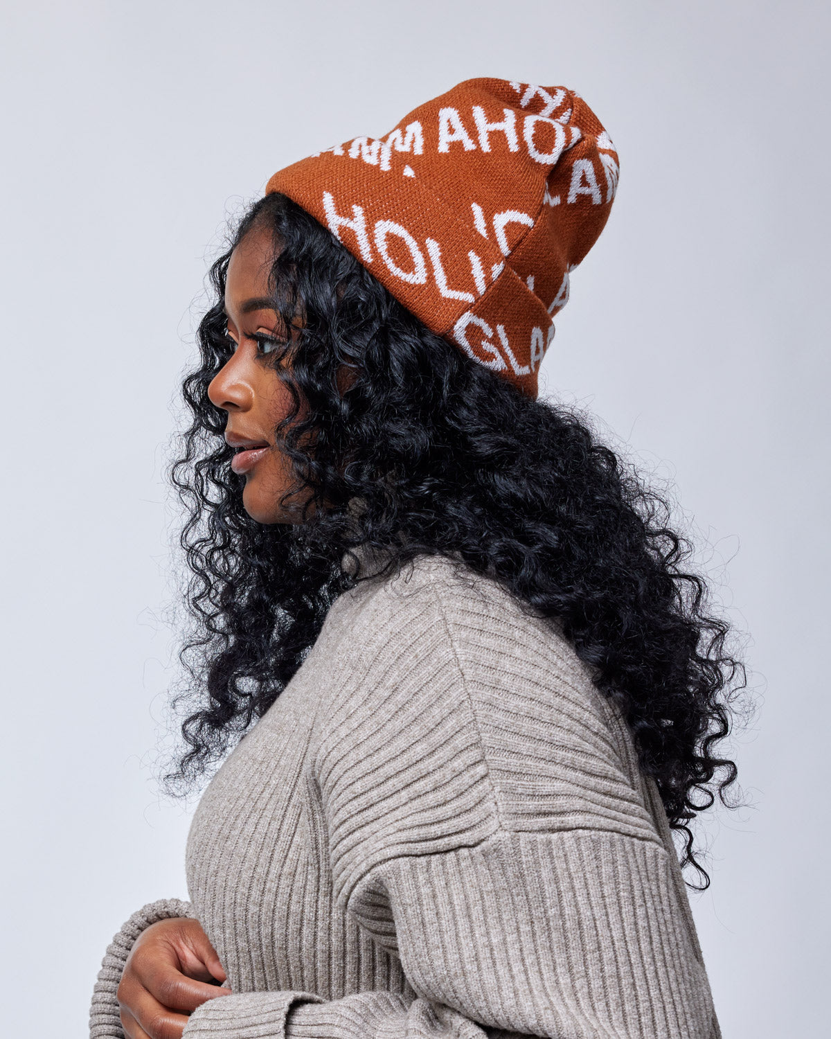 Woman wearing a brown Monogram Beanie with white lettering, paired with a beige ribbed sweater, against a plain background.