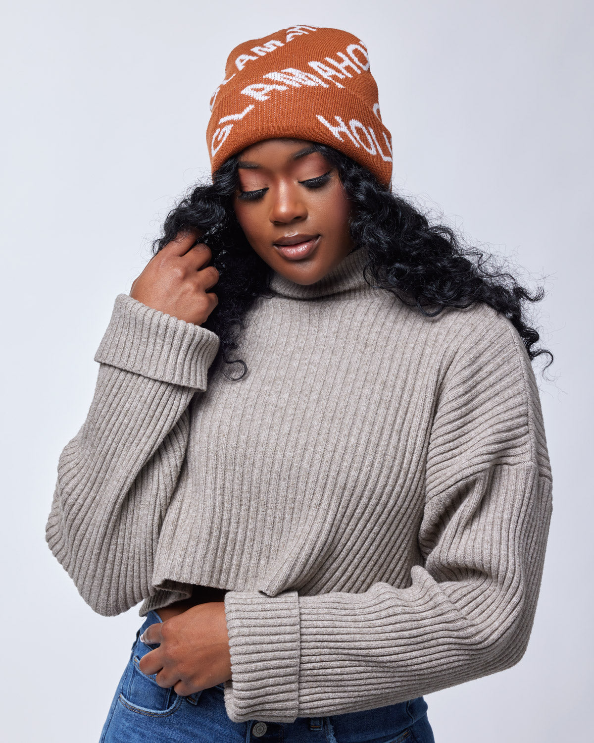 Woman wearing a brown monogram beanie with white text, beige ribbed sweater, and blue jeans against a plain background.