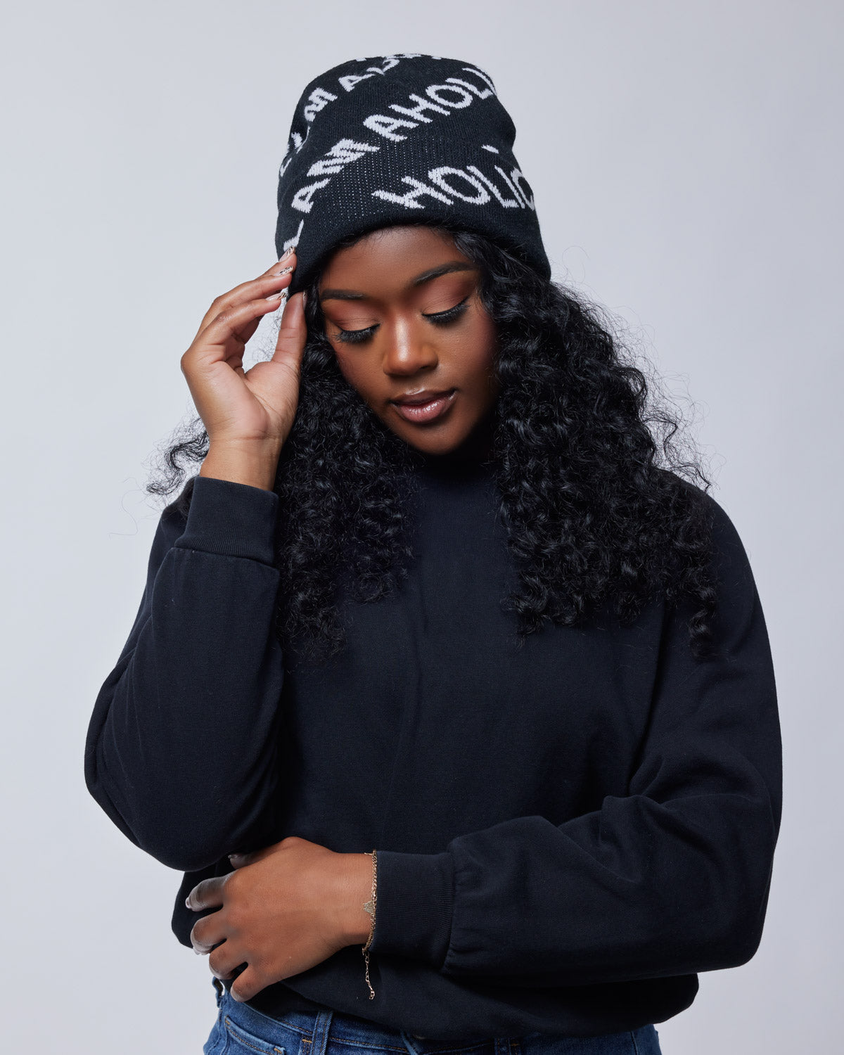Black Monogram Beanie with white logo text, worn by a woman with curly hair, dressed in a black sweatshirt against a plain background.
