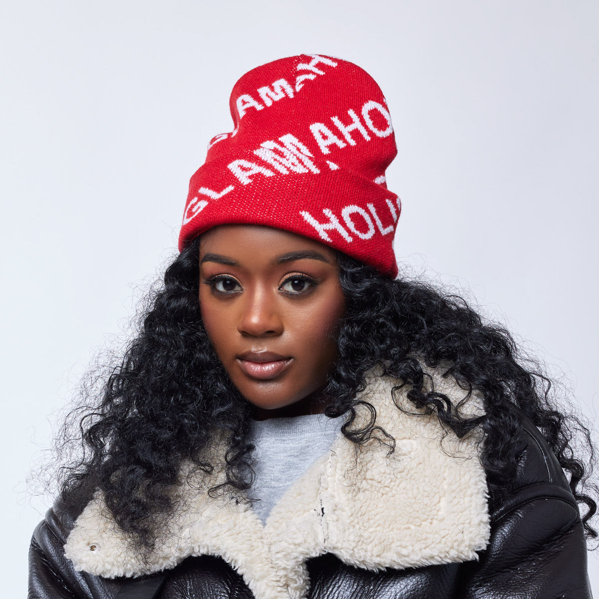 Red monogram beanie with white lettering, worn by a woman with curly black hair, styled with a shearling-lined leather jacket.