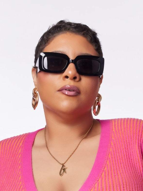 Stylish woman wearing oversized black sunglasses, gold hoop earrings, and a pink sweater, showcasing a chic look.