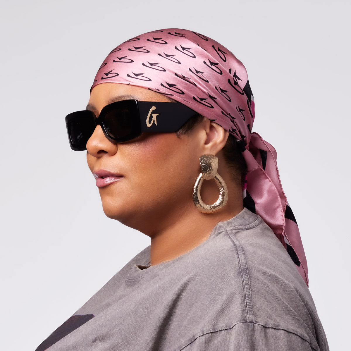 Woman wearing a pink monogram silk scarf, oversized sunglasses, and statement earrings, styled with a casual gray top.