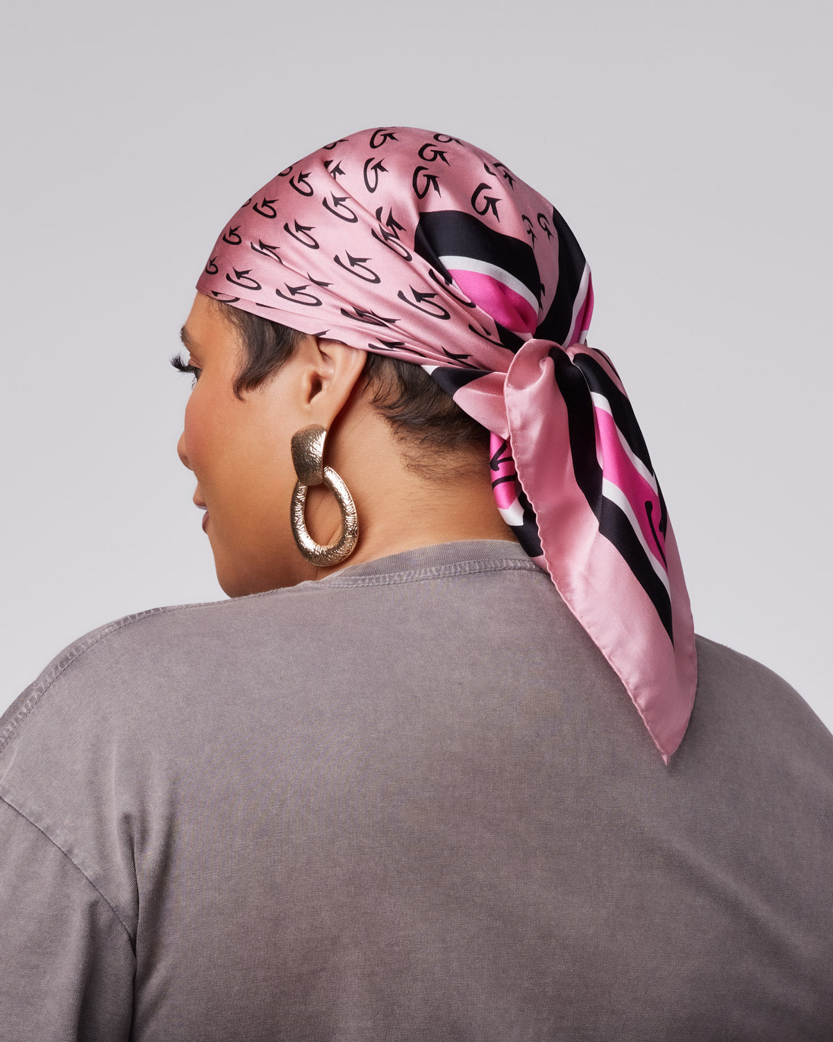 Medium pink silk scarf featuring a monogram design, styled as a head wrap on a model with large earrings.