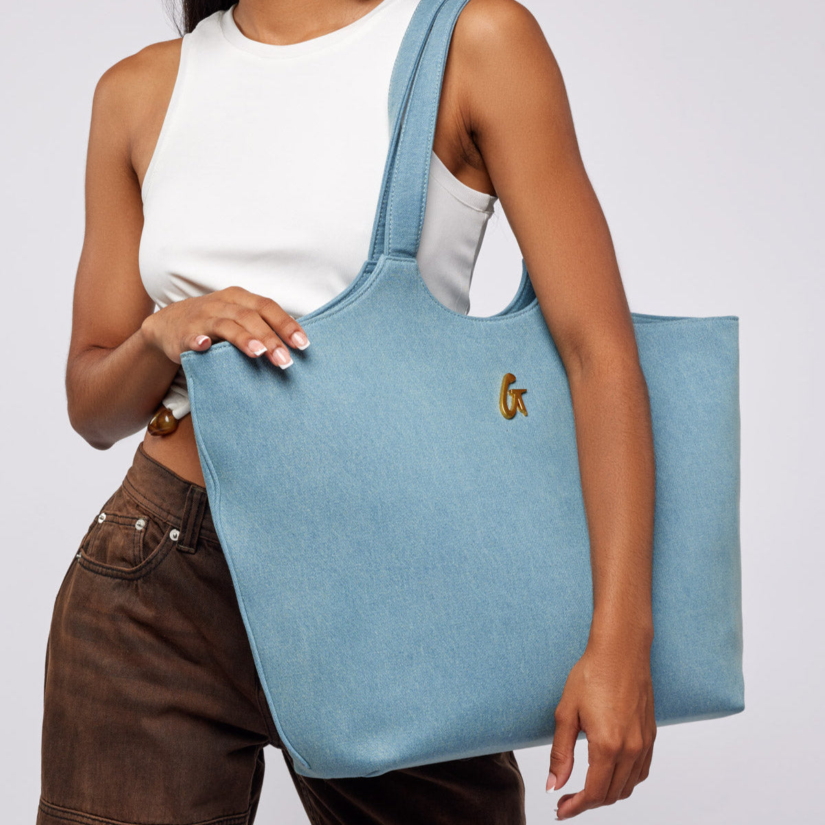 Light blue denim maxi tote bag with a gold logo, held by a model wearing a white tank top and brown pants.