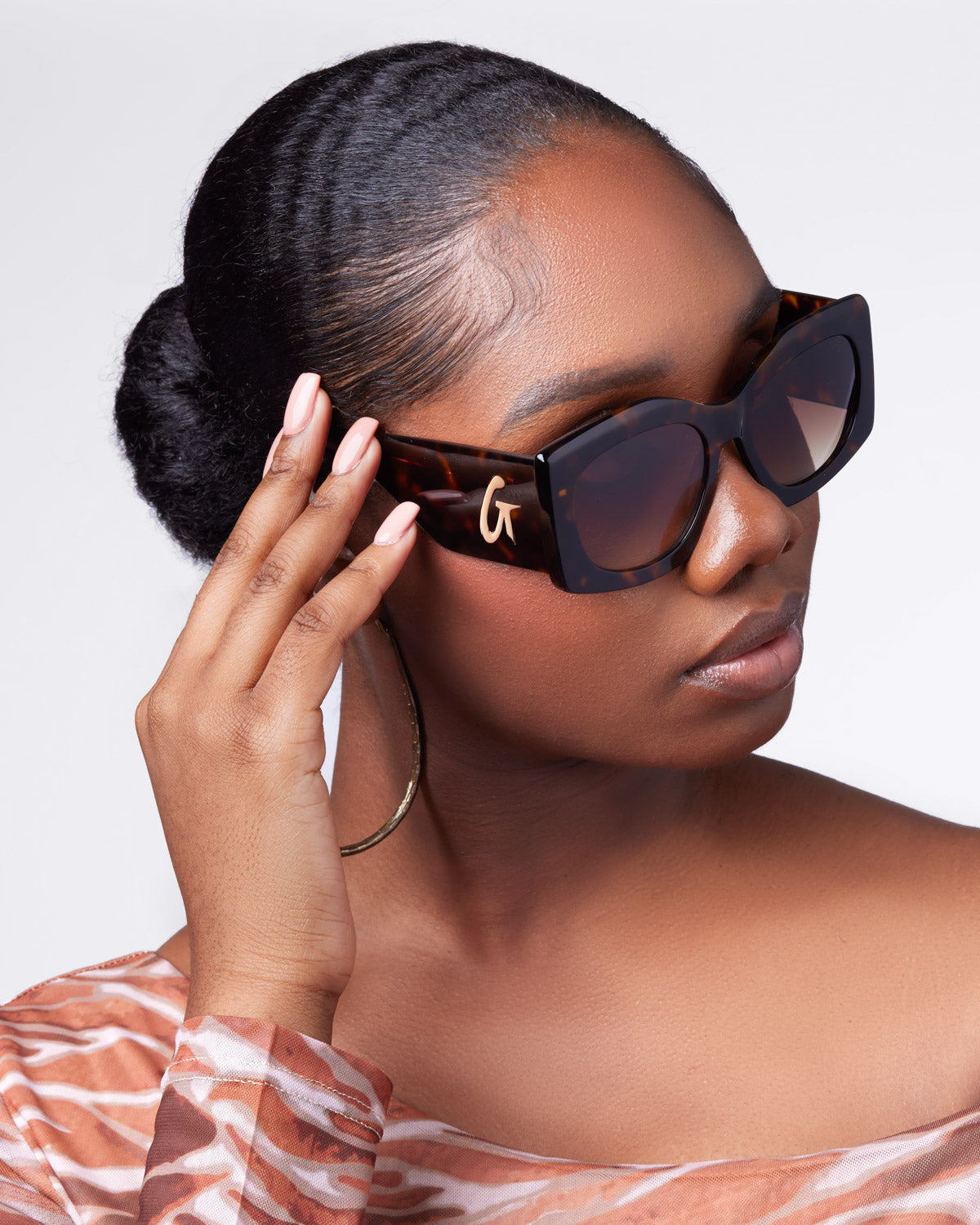 A woman wearing oversized tortoise sunglasses, gently touching the frame, with a stylish off-shoulder top and elegant hairstyle.