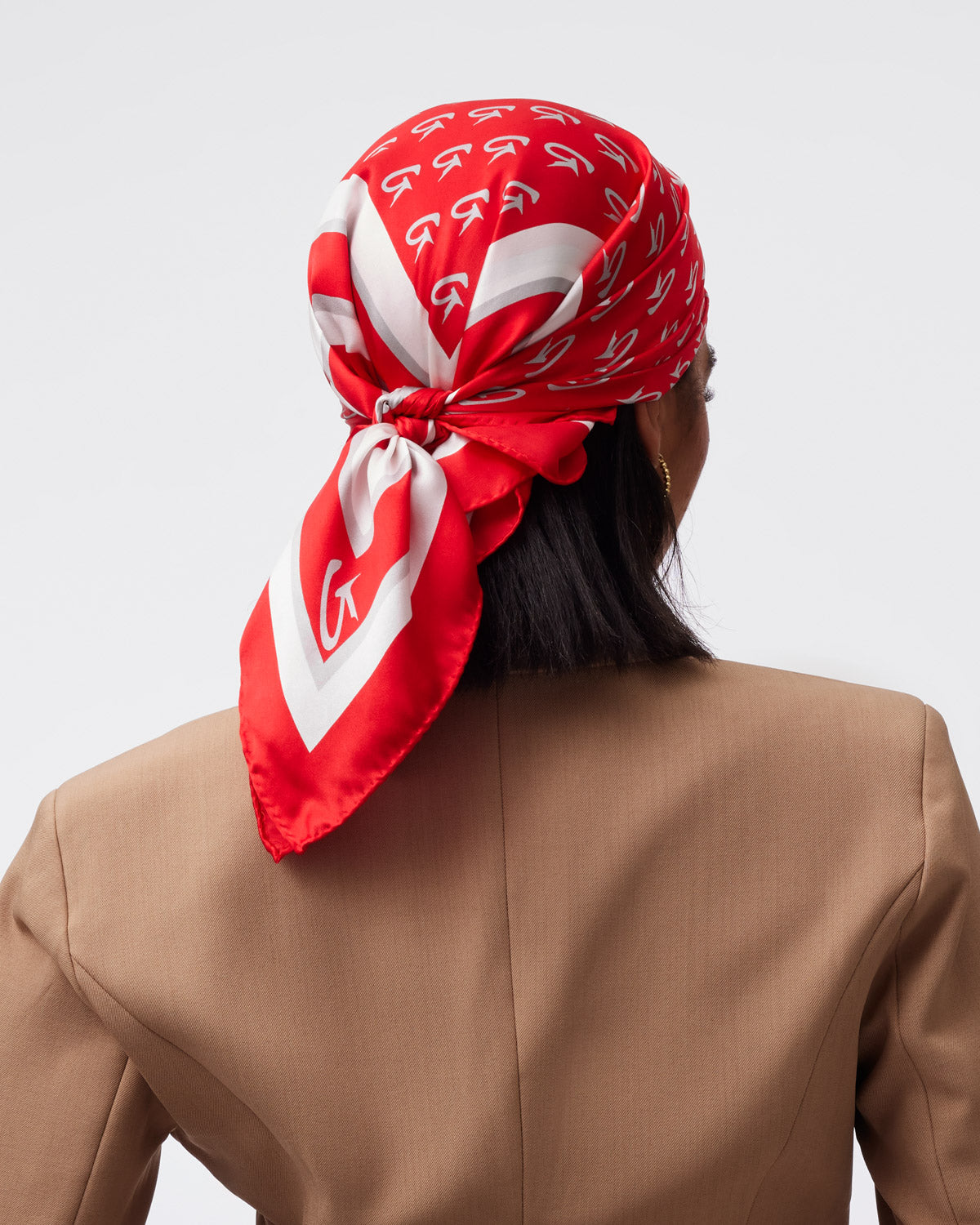 Medium red silk scarf featuring a monogram pattern, elegantly tied at the back of a model's head, complementing a beige blazer.