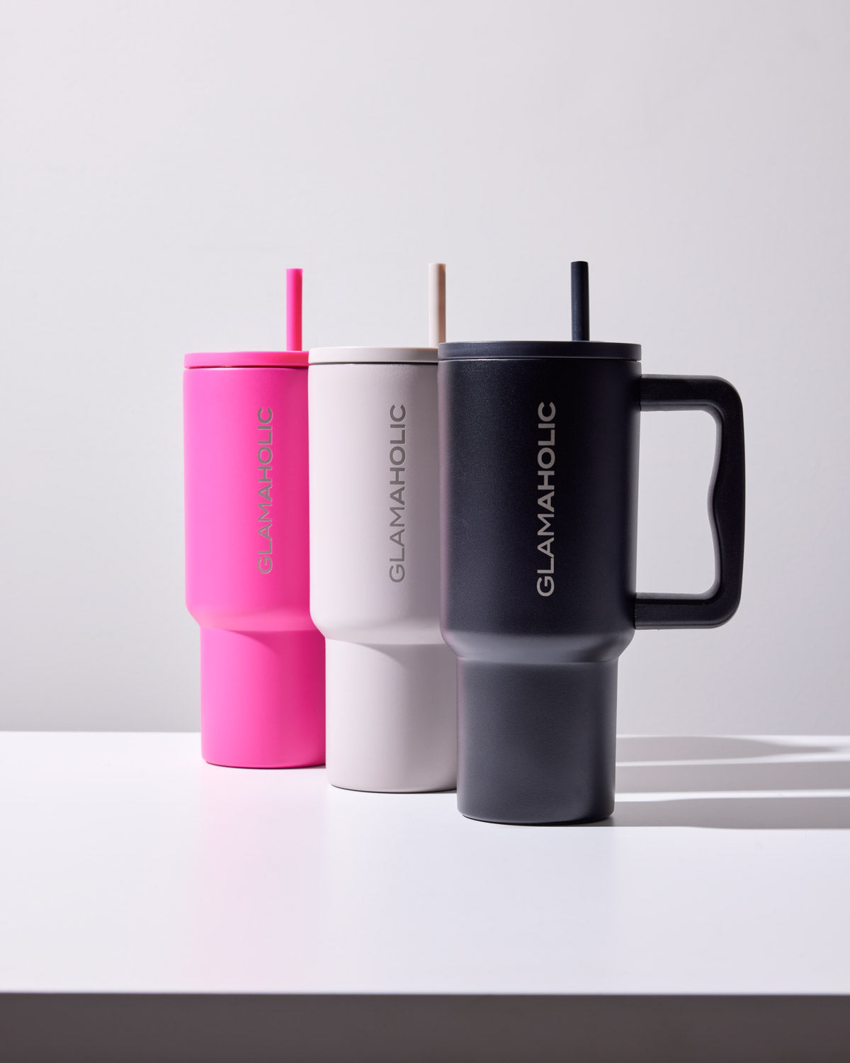 Set of three stylish travel mugs in pink, white, and black with "GLAMAHOLIC" logo, matching straws, and ergonomic handles.