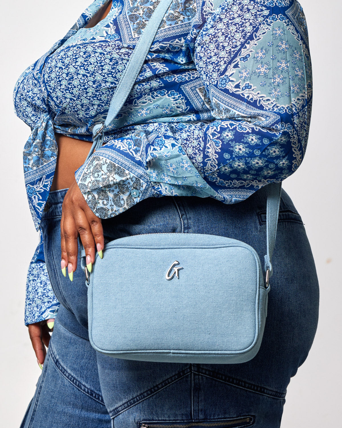 A light blue denim camera bag with a silver logo, worn crossbody by a person in a blue patterned shirt and jeans.