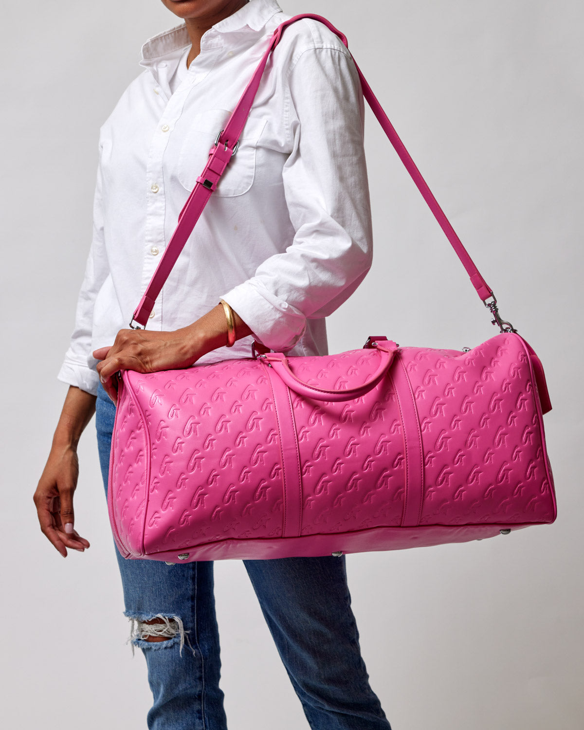 Hot pink monogram duffle bag with embossed logo pattern, carried by a woman in a white shirt and ripped jeans.