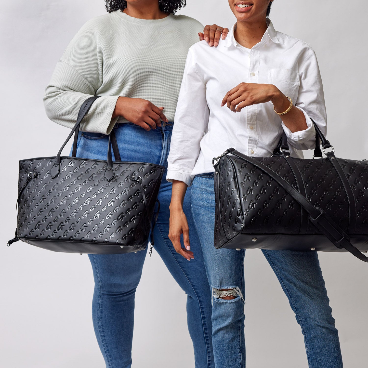 Two women hold black Monogram Glam Totes with pink accents, featuring a repeating logo pattern, against a plain background.