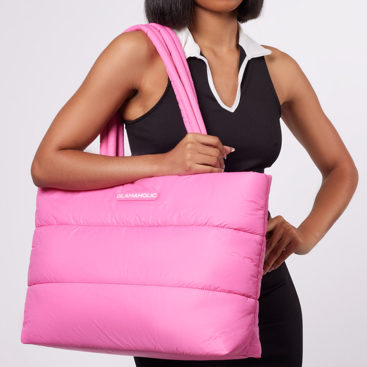 Pink quilted tote bag with "GLAMAHOLIC" logo, carried over the shoulder by a woman in a black sleeveless top with white co...