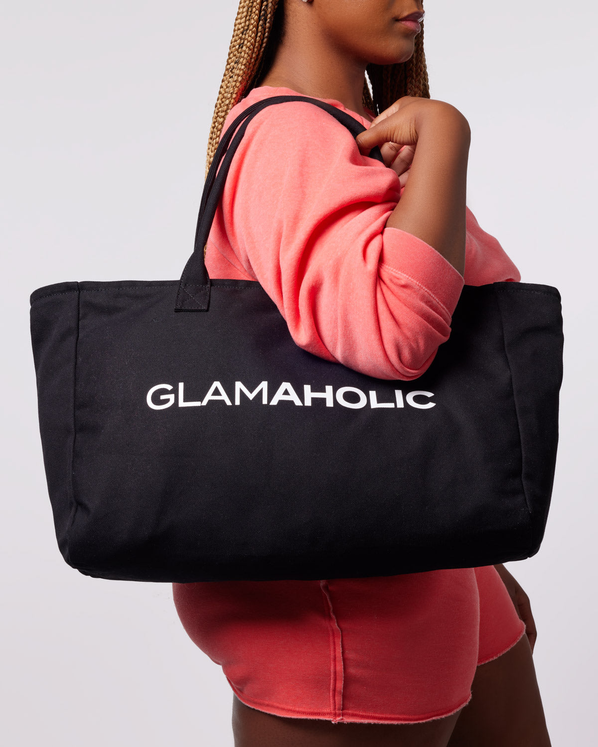 Black athletic canvas tote bag featuring the word "GLAMAHOLIC" in bold white letters, held by a model in a pink outfit.
