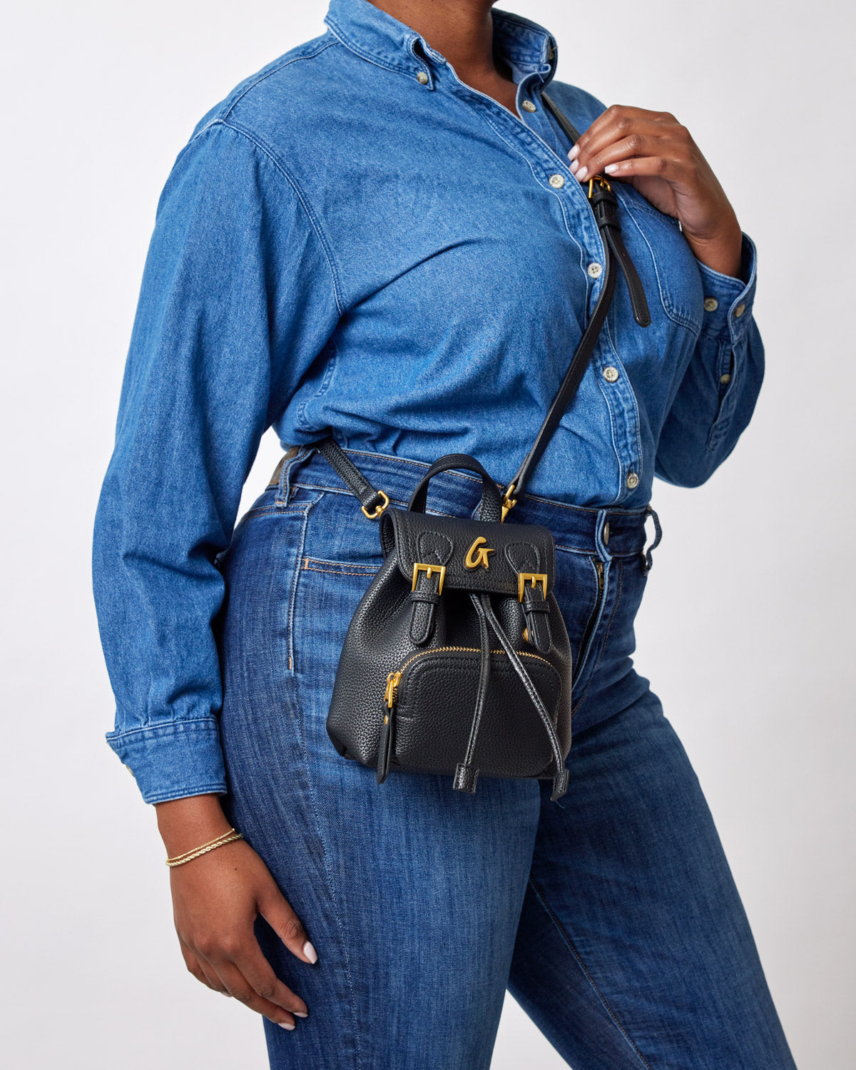 Black mini pebble crossbody backpack with gold hardware, worn by a person in a denim outfit.