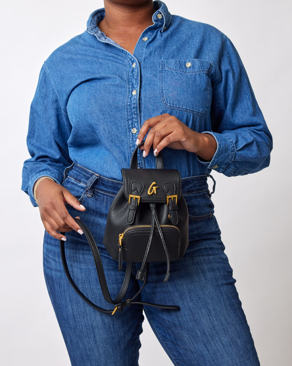 Mini black pebble crossbody backpack with gold hardware, worn by a person in a denim shirt and jeans.