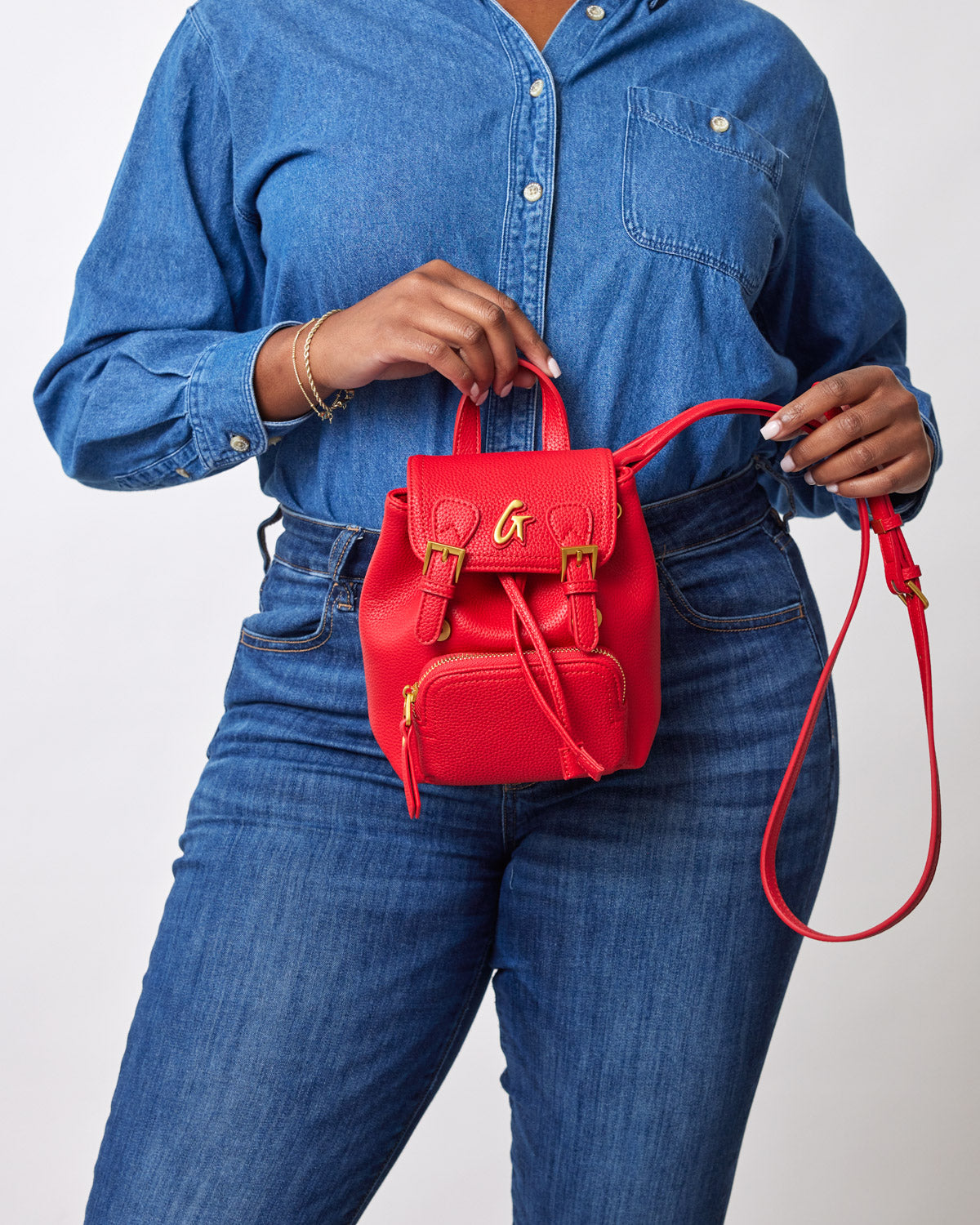 Red mini pebble crossbody backpack with gold hardware, front zipper pocket, and adjustable strap, worn by a person in denim.