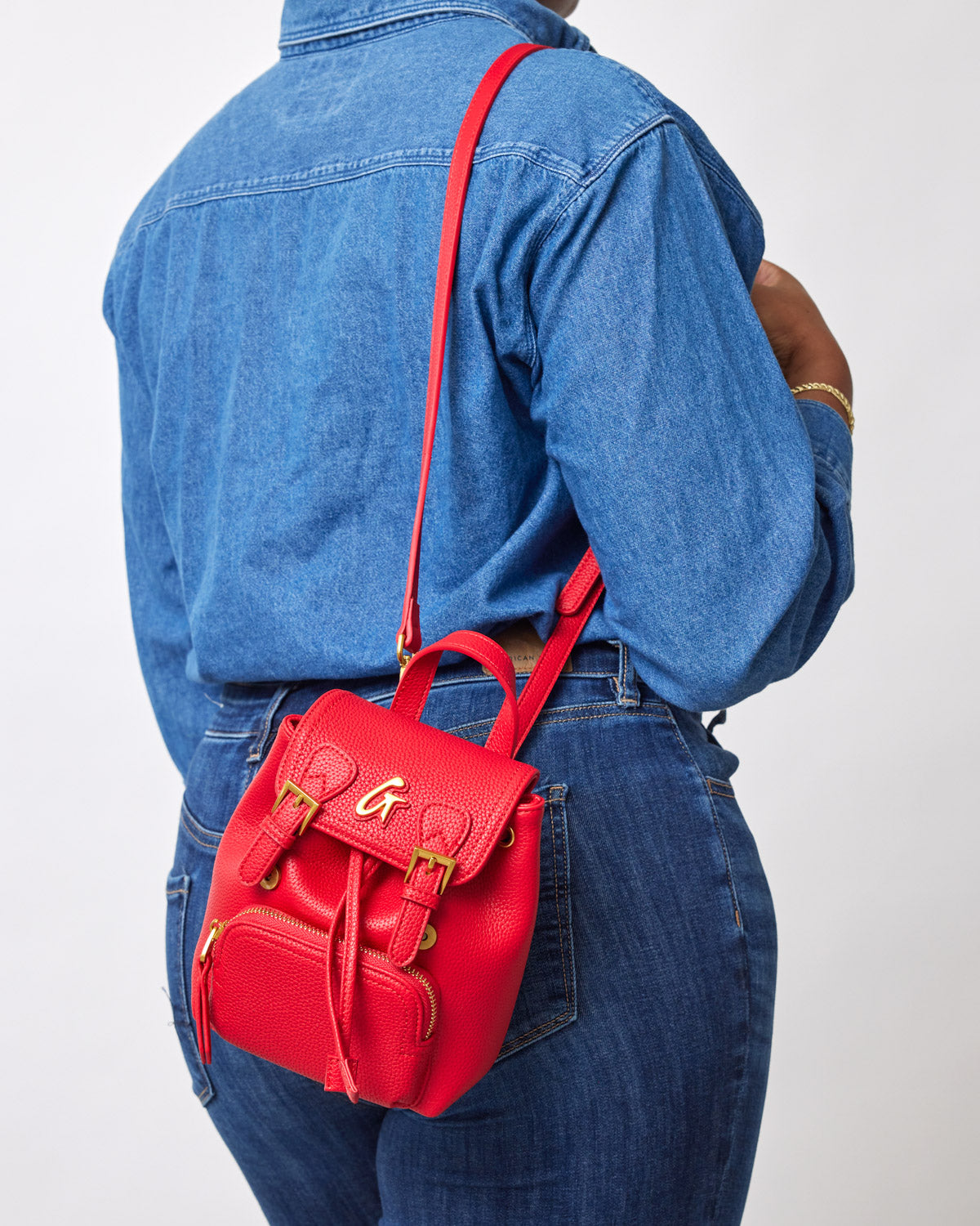 Red mini pebble crossbody backpack with gold hardware, worn over a blue denim shirt and jeans.