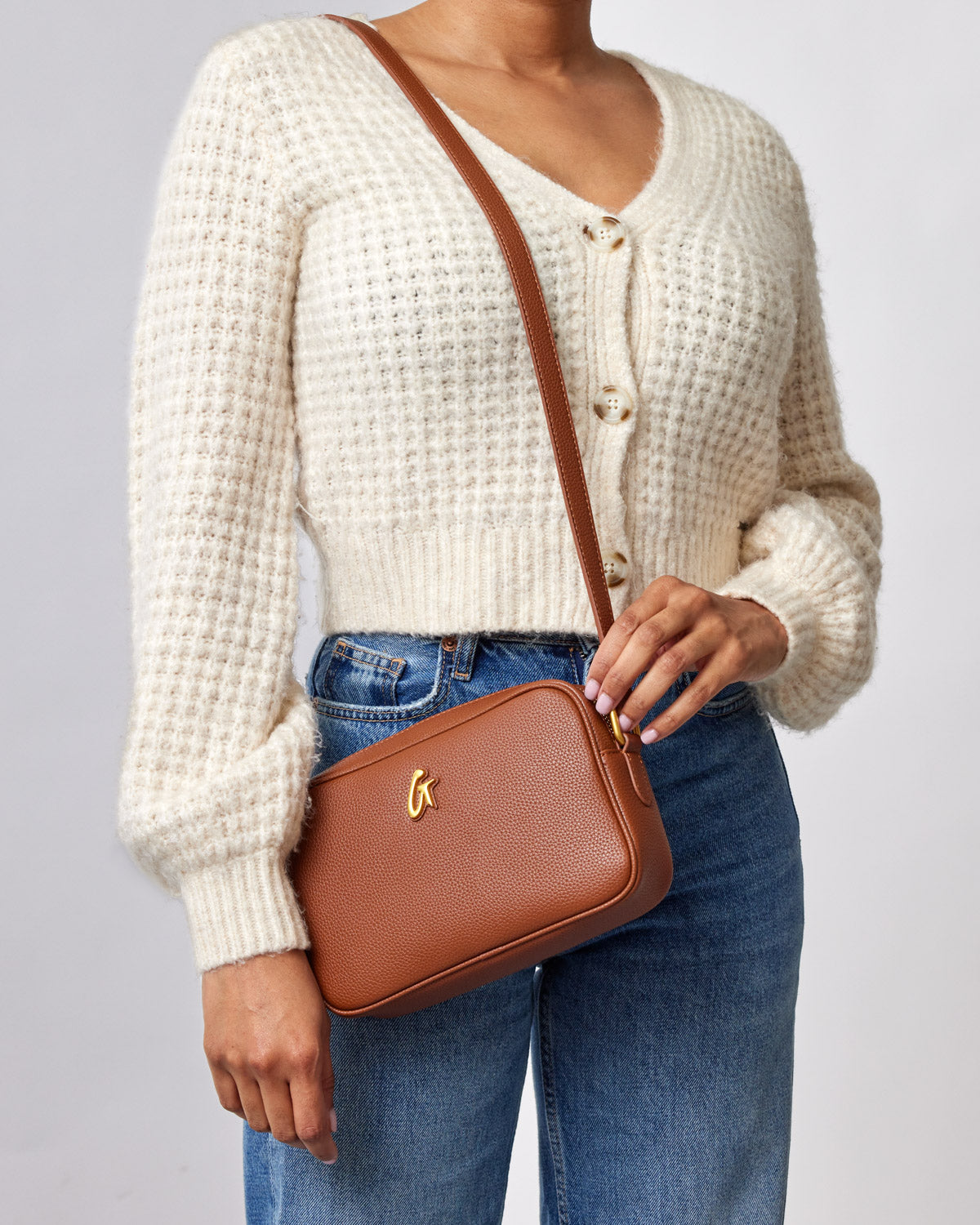 Brown pebble-textured camera bag with a gold logo, worn crossbody over a cream knit sweater and blue jeans.
