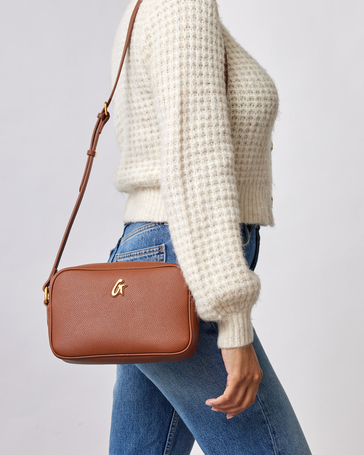 Brown pebble-textured camera bag with a gold logo, worn crossbody over a cream sweater and blue jeans.