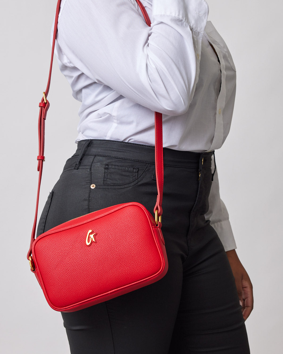 Red pebble-textured camera bag with gold logo, worn crossbody over black pants and white shirt.
