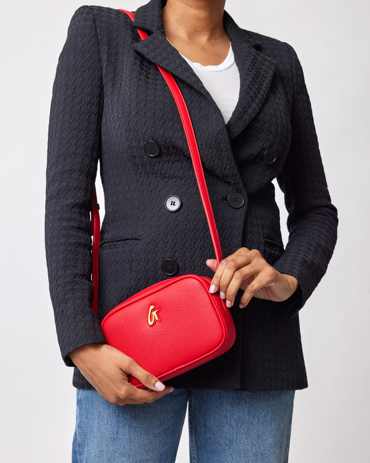 Red MINI PEBBLE CAMERA BAG with a gold logo, worn crossbody over a black blazer, held by a person in jeans.