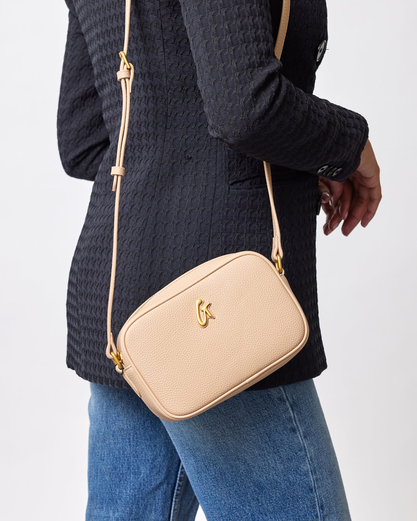 A beige pebble-textured camera bag with a gold logo and adjustable strap, worn crossbody over a black textured blazer.
