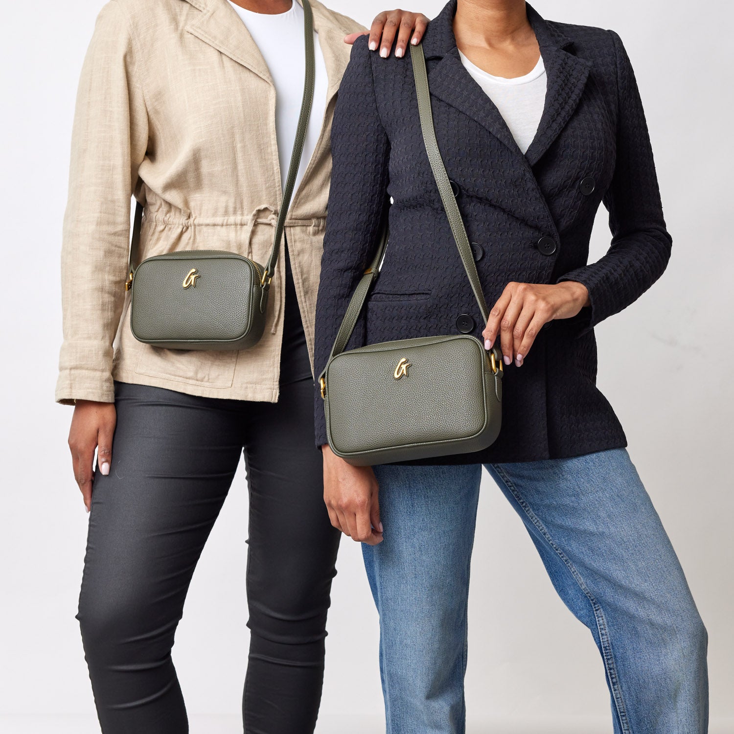 Two women display olive green pebble-textured camera bags with gold logo, wearing casual and blazer outfits.