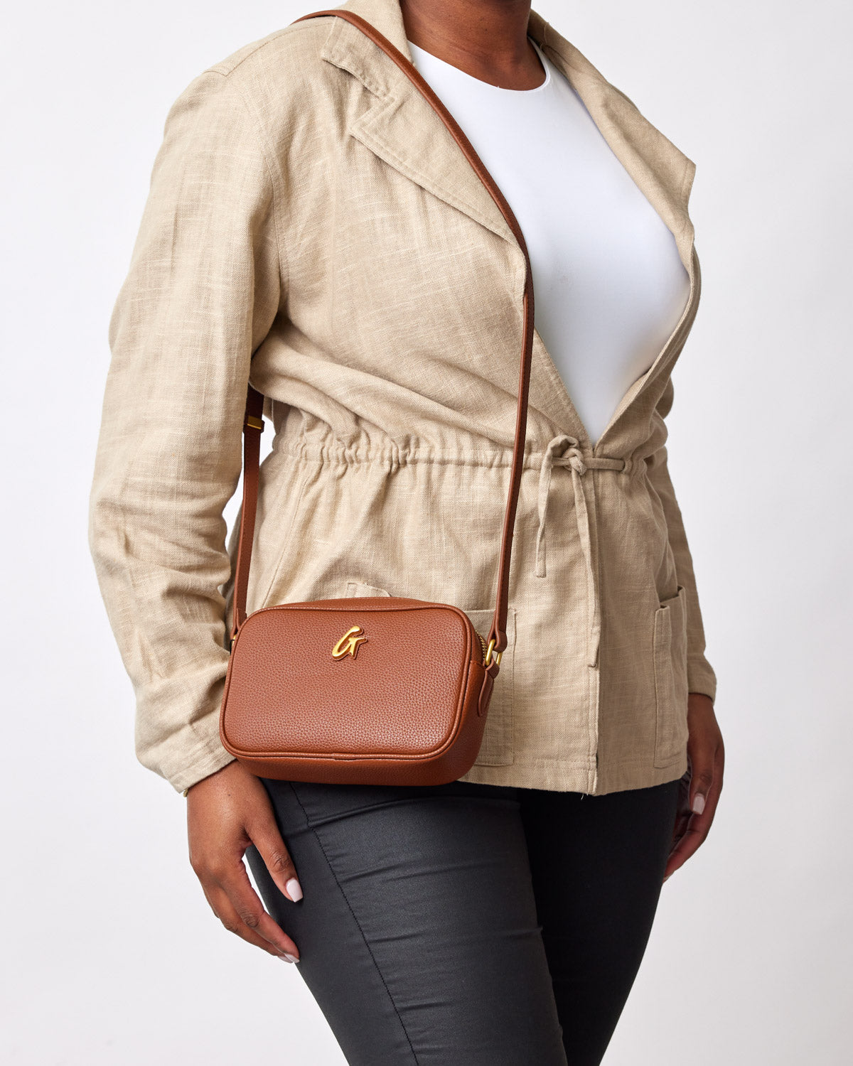 Mini Pebble Camera Bag in brown with gold logo, worn crossbody over beige jacket and black pants.