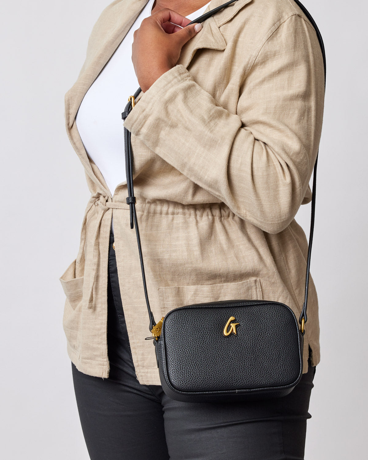 Black mini pebble camera bag with a gold "G" logo, worn crossbody by a person in beige blazer and black pants.