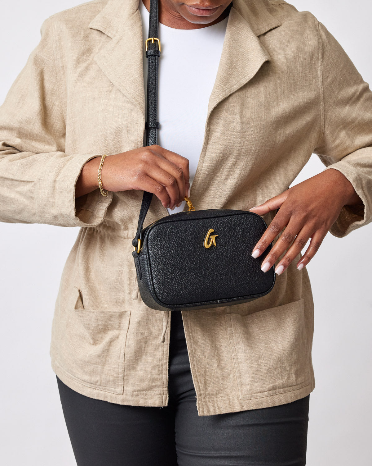 Small black pebble-textured camera bag with gold logo, worn crossbody by a person in beige jacket and black pants.
