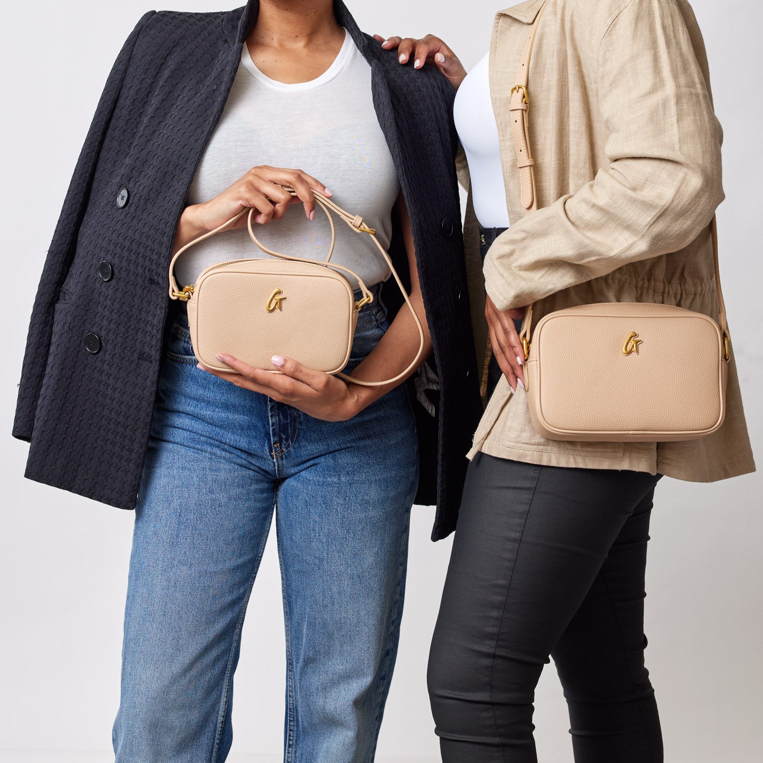 Two women hold beige Pebble Camera Bags, one with a short handle and the other with a crossbody strap, against a plain background.