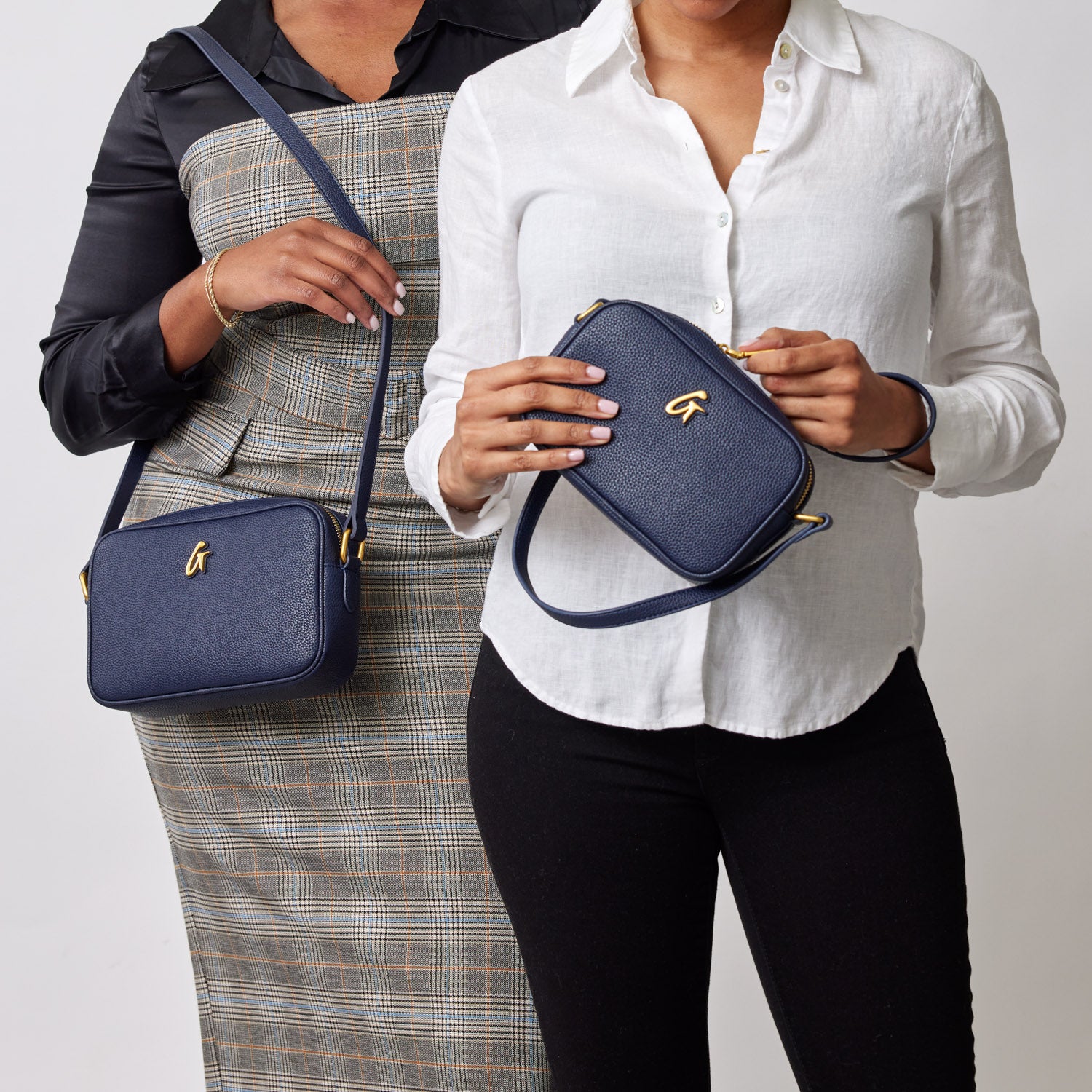 Two women hold navy mini pebble camera bags with gold logo and adjustable straps, showcasing stylish accessories.