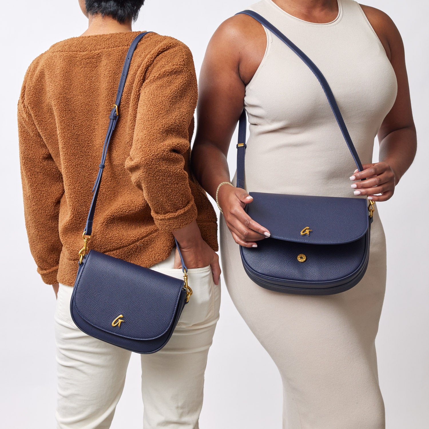 Two women showcase navy medium pebble shoulder bags with gold accents, styled with casual outfits against a plain background.