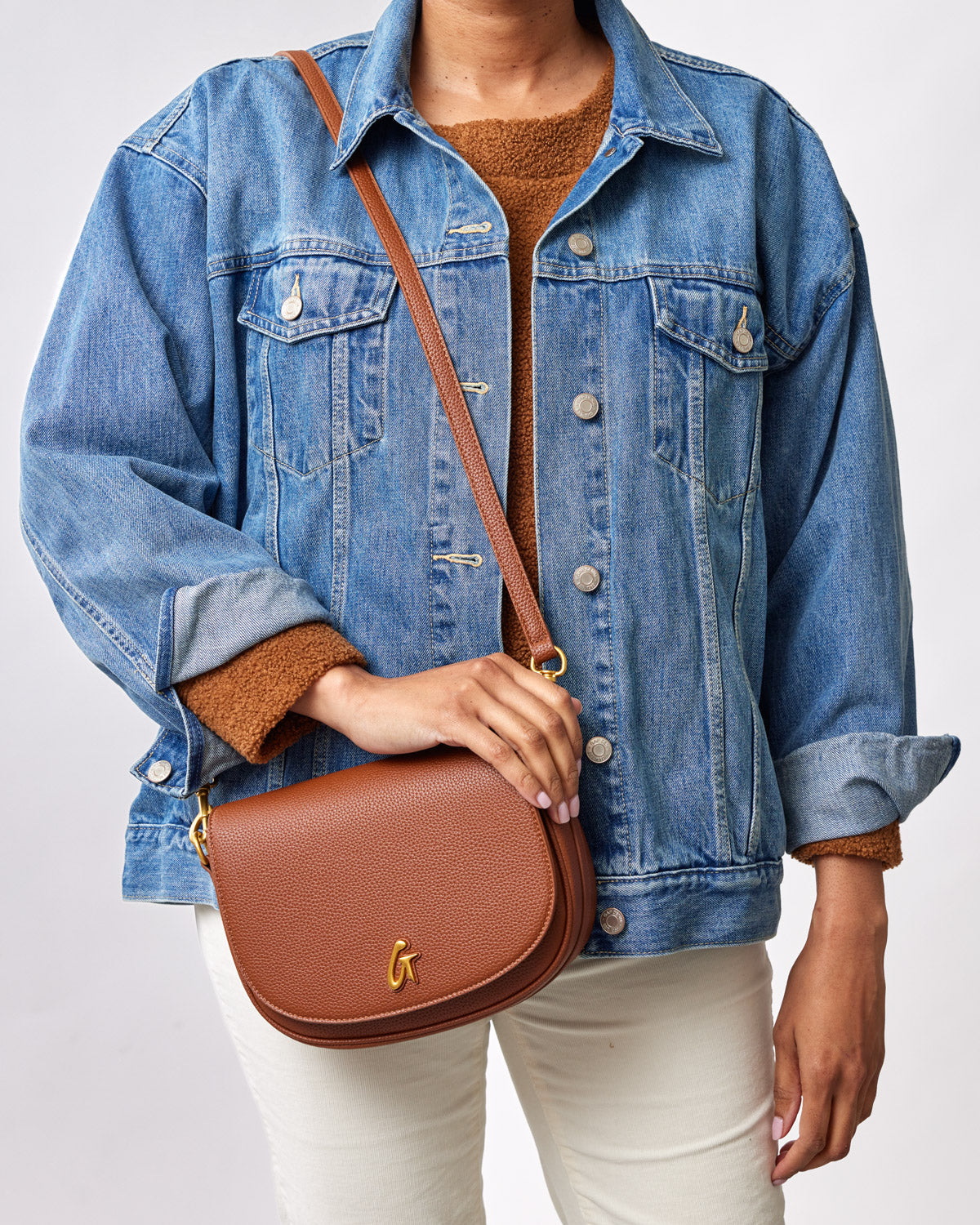 Medium brown pebble shoulder bag with a gold logo, worn crossbody over a denim jacket and beige pants.