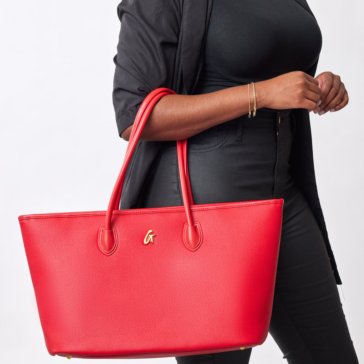 Red pebble tote bag with dual handles, gold logo, and spacious design, carried by a woman dressed in black.