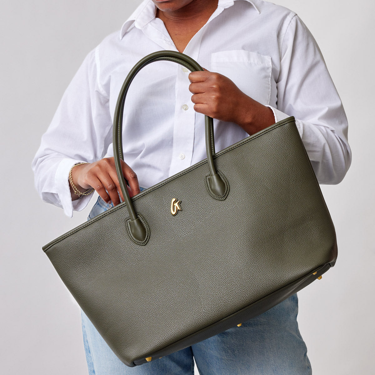 Olive green Pebble Tote Bag with dual handles, gold logo, textured surface, held by a person wearing a white shirt and jeans.