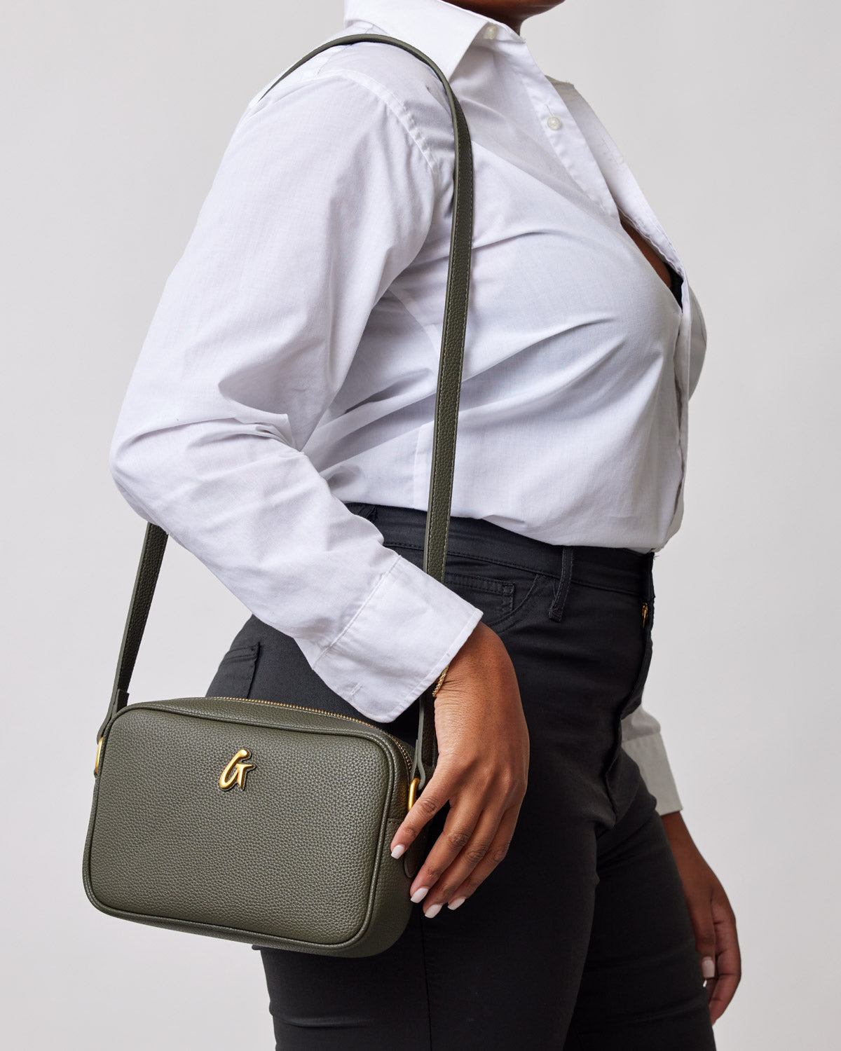 Olive green textured handbag with gold hardware and a 'G' logo, worn crossbody with a white button-up shirt and black pants.