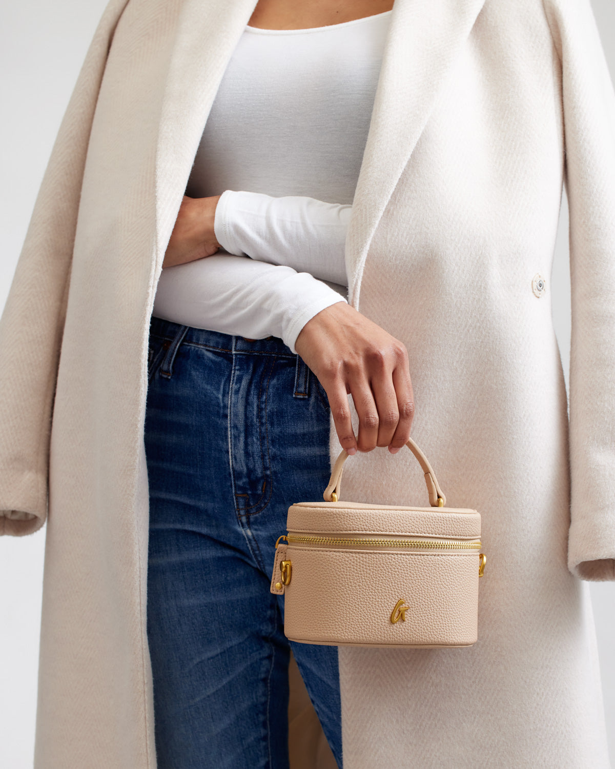 A woman holding a nude-colored mini pebble vanity bag with a top handle and gold zipper, dressed in a white top, jeans, and a cream coat.